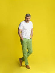 Middle aged handsome grey hair man posing with hands in pockets and crossed legs wearing white t-shirt and green jeans isolated on yellow background. Fit, muscular middle aged man