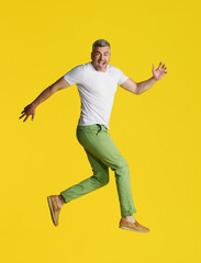 Fototapeta na wymiar Handsome man with grey hair jump in the air turned sideways wearing white t-shirt and green jeans isolated on yellow background. Excited, happy fit, muscular middle aged man