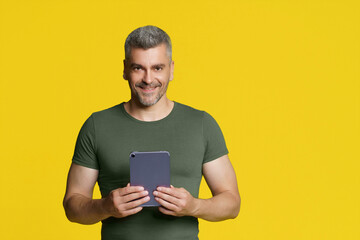 Smart middle aged handsome man holding digital tablet in hands smiling looking at camera isolated on yellow background. Happy freelancer man with tablet. Using modern technologies grey haired man