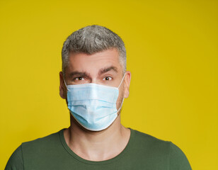 Confused, suspicious handsome mature grey haired man wearing facial medical mask. Mature caucasian man in casual and medical mask isolated on yellow background. Pandemic concept