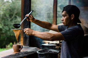Young brown man serving coffee with a strainer in a traditional Mexican kitchen.