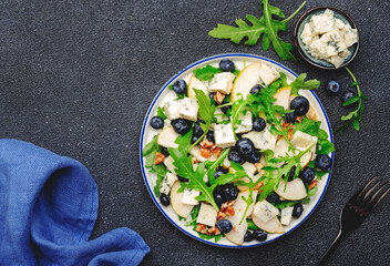 Delicious arugula salad with pears, blueberries, roquefort cheese and walnuts. Black kitchen table background, top view