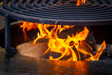A wooden log burns brightly in a wood-fired grill. Circle wood-fired grill with steel grate for outdoor cooking