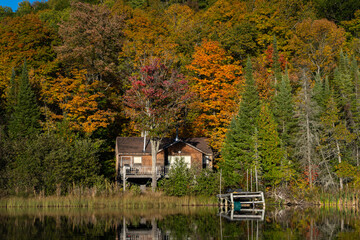 An old cabin sits nestled between a lake and the colourful fall foliage.