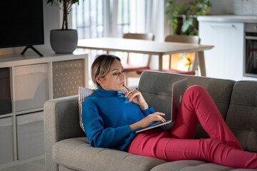 Focused Caucasian woman with laptop lies on sofa in living room of home. Pensive girl writer in comfortable environment writes own novel or book for Internet subscribers posing in lifestyle style