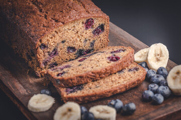 Loaf of fresh baked homemade blueberry banana bread and slices of fruit on a wooden cutting board