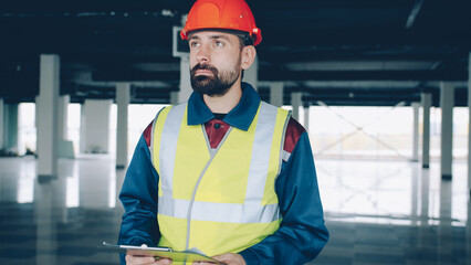 Construction inspector wearing uniform is checking papers and studying industrial building inside. Supervision and hse inspection concept.