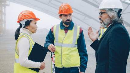 Builders wearing uniform are talking at construction site while architect in suit and helmet is coming discussing project gesturing and looking around - 536220552
