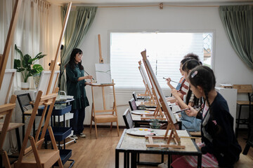A female Asian teacher teaches and demonstrates to the children on acrylic color picture painting on canvas in art classroom, creatively learning with skill at the elementary school studio education.
