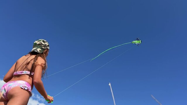 Low angle view of standing little redhead girl with hat on summer holidays flying green kite with long tail high in clear blue sky by holding and pulling handles and strings