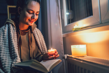 Teenage girl sits under blanket near heating radiator with candles and read book .Rising costs in...