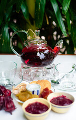 Hibiscus tea in a glass teapot with cups and crystalized fruits and jam on a table