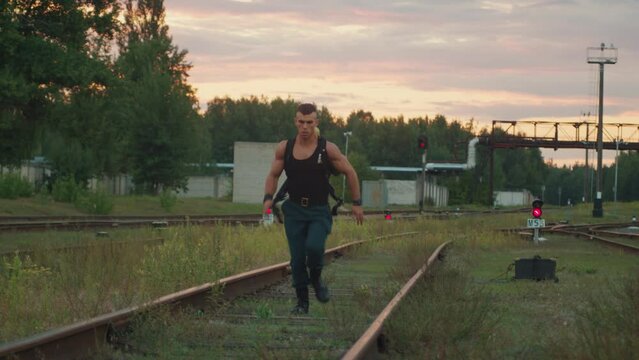 Muscular man training and run on rails with heavy load on his back against the backdrop of pipeline and semaphores with red light