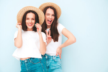 Two young beautiful smiling hipster female in trendy same summer white t-shirt and jeans clothes. carefree women posing near blue wall in studio. Cheerful and positive models in hats