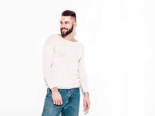 Fototapeta premium Portrait of handsome smiling stylish hipster lambersexual model.Man dressed in white sweater and jeans. Fashion male posing near wall in studio. Isolated
