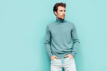 Tuinposter Portrait of handsome confident  model. Sexy stylish man dressed in  sweater and jeans. Fashion hipster male with curly hairstyle posing near blue wall in studio. Isolated © halayalex