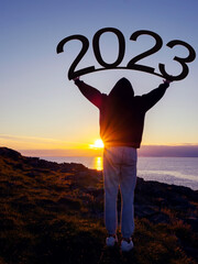 Silhouette of a teenager girl holding sign 2023 above her head. Sunrise sun in the background....