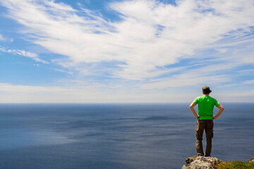 Man standing on an edge of a rock and looking at vast ocean and blue cloudy sky in the background. Back to viewer. Reaching you goals and self belief concept. Copy space. Travel and tourism concept.