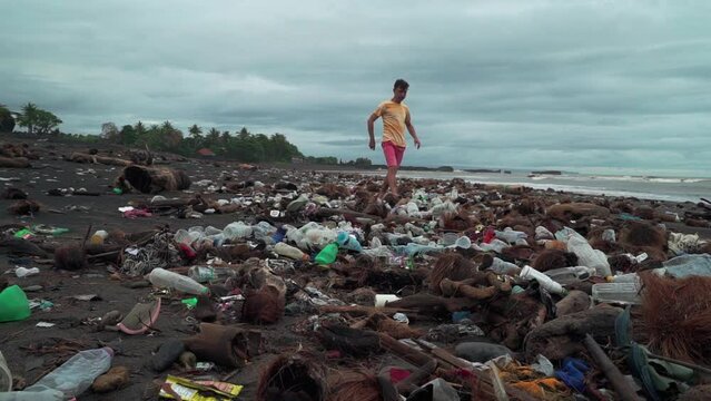 Garbage and plastic waste on the beach against a gloomy sky. A lone man tourist walks along the muddy shore, stepping over trash. Environmental pollution ecological problem.