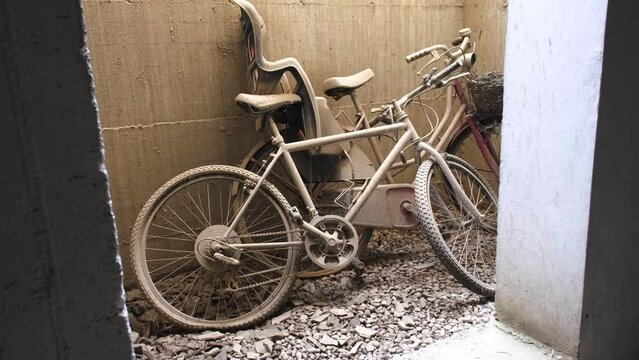 Two abandoned bikes with flat tires left leaning on a sandy wall, on rubble covered ground. With a child seat, covered in dirt
