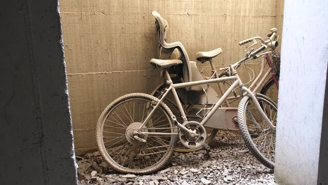 Two abandoned bikes with flat tires left leaning on a sandy wall, on rubble covered ground. With a child bike seat, covered in dirt