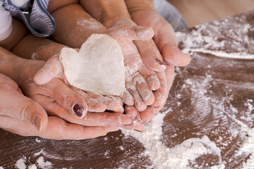 Family in kitchen, close-up palm from above lies a heart-shaped dough on a wooden table with flour,...