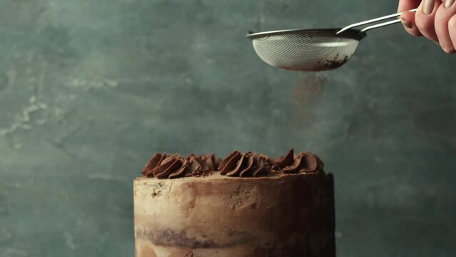 Pastry Chef's hand sprinkles chocolate cake with cocoa powder in a slow motion. Sponge cake with brown frosting decorated with chocolate buttercream topping on rustic concrete background. 