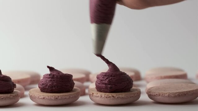 Process of making French dessert macaroon. Close up of Chef using pastry bag to squeeze creamy berry filling on pink macaroons halves.  High quality 4k footage. 