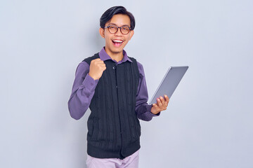 Smiling young Asian man in casual shirt and vest holding tablet pc computer and making winner gesture isolated on white background. People lifestyle concept