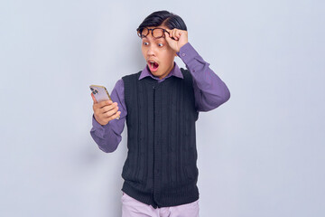 Surprised young Asian man in casual shirt using smartphone and taking off glasses isolated on white background
