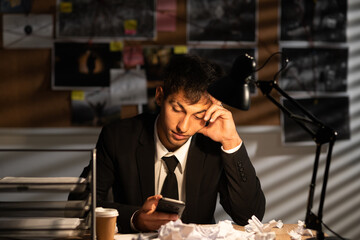Worried frustrated detective in dark suit sitting at an office desk full with papers being...