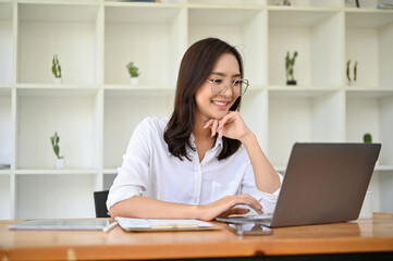 Fototapeta na wymiar Attractive Asian businesswoman in eyeglasses looking at laptop screen, hand on chin