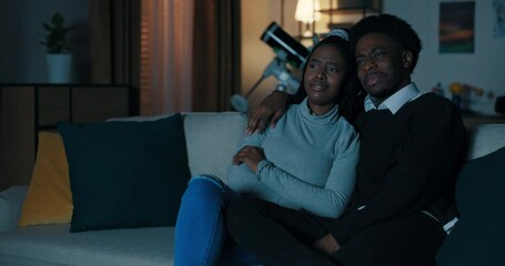 Young man of African origin asked attractive girl on date. Couple in love is sitting on sofa in the living room and watching movie. The guy gently hugged the girl. They discuss the movie and smile.