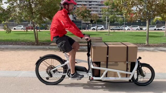 young messenger with red clothes and helmet riding a cargo bike to make a shipment on a bike lane in a city, horizontal video