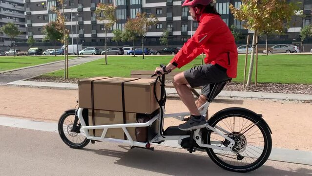 young messenger with red clothes and helmet riding a cargo bike to make a shipment on a bike lane in a city, horizontal video
