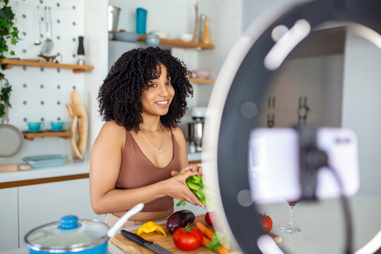 Happy African American woman smiling and demonstrating ripe vegetables while shooting video for cooking vlog in kitchen at home