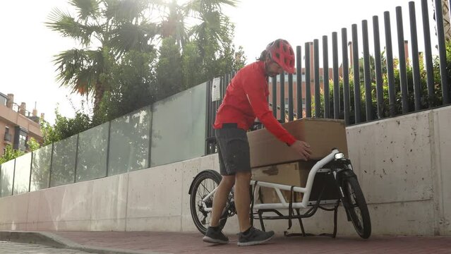 young courier in red clothes and helmet riding cargo bike carrying a large package to be shipped in the city, horizontal video