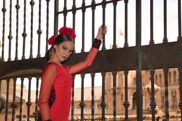 Portrait of young teenage woman in red dance suit with red carnations in her hair doing flamenco poses holding on to a fence. Flamenco concept, dance, art, typical Spanish dance.