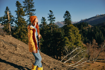 Naklejka premium Woman traveling to the mountains in the fall on a nature hike smile and happiness in a yellow cape with red hair full-length stands against the backdrop of trees and mountains