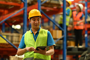 Asian male manager in hardhat and reflective jacket using digital tablet, checking inventory production stock control in warehouse