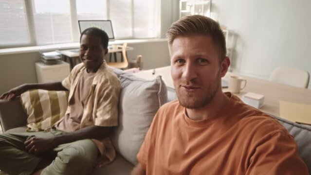 Handheld pov slowmo of two diverse male friends smoking electronic devices at home, chilling on sofa and speaking at camera