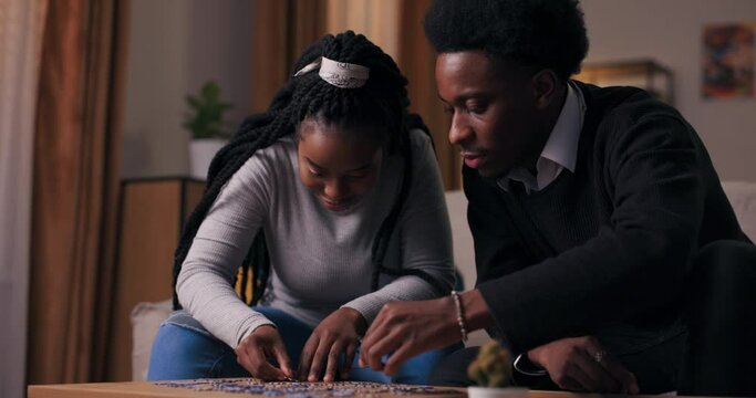Brother and sister of African appearance like to spend time together and do puzzles. They concentrate on creating picture. Girl with long hair and guy in black suit are talking and having fun.