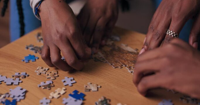 Close-up of hands of people of African appearance making puzzles. The boy has silver ring on finger. Friends collect puzzles on wooden table. The picture is dominated by blue and brown colors.