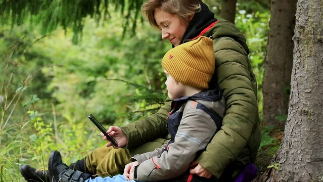 Joyful happy traveling mother and son using smartphone mobile resting in mountains forest near the lake enjoying beautiful autumn nature. Internet connecting 5g. Young family lifestyle.