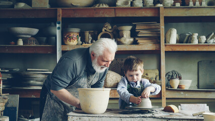 Curious little boy is learning pottery from his experienced grandfather in small home studio. Child is forming clay to make pot on potter's wheel, his granddad is helping him. - obrazy, fototapety, plakaty