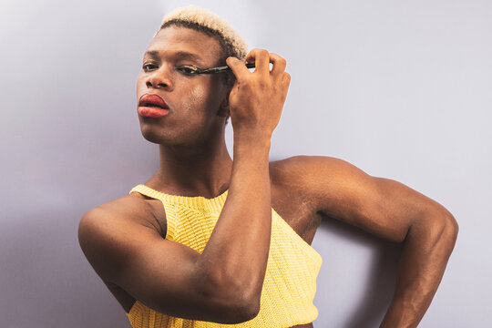 An androgynous black man posing against a purple studio background while applying eye makeup.
