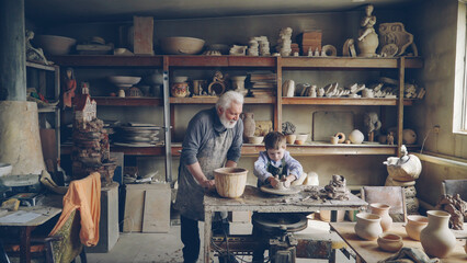 Experienced potter loving prandparent is teaching his young grandchild pottery in small home workshop. Boy is working with throwing wheel, grandpa is talking to him. - obrazy, fototapety, plakaty