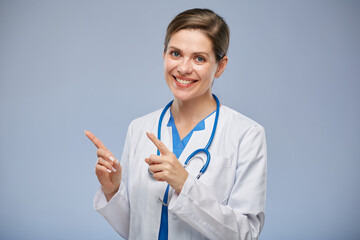 Smiling doctor woman in white lab coat pointing with two fingers at left side, isolated portrait.