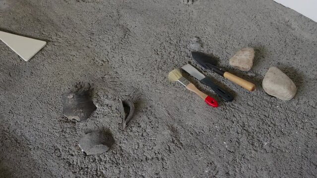 Historical excavations, brush and spatula tools lie on the sand, a broken clay pot.
