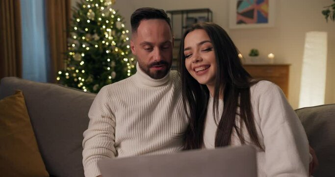 Winter evening. Young married couple is sitting on sofa and looking at children's photos on laptop. Girl with long black hair shows school photos to husband. They are having fun, talking and smiling.
